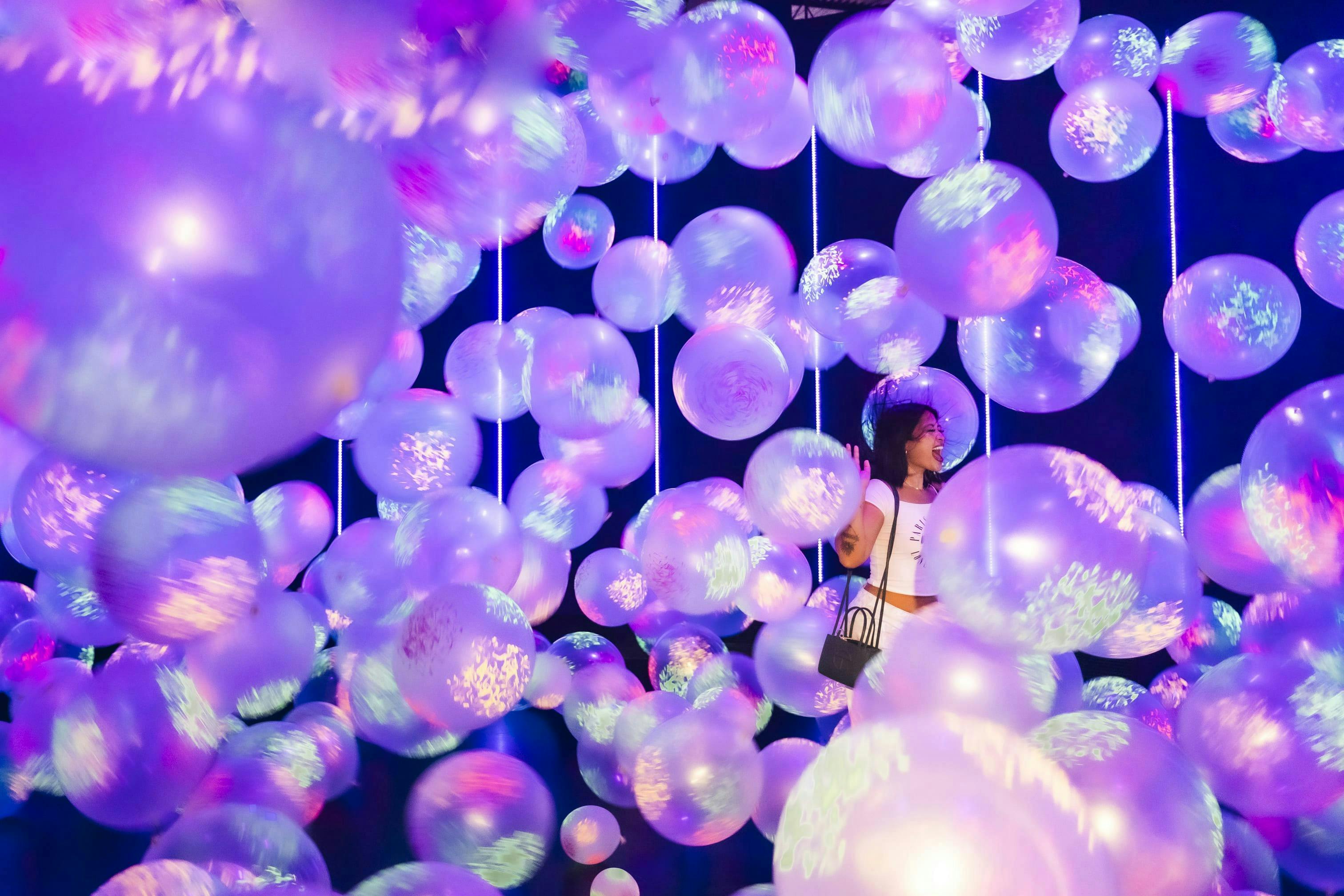 紫色の照明の下、輝く半透明の風船に囲まれた女性が立っており、活気に満ちた没入型のシーンを作り出しています。