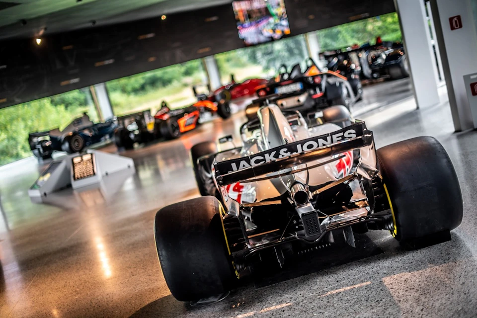 A Formula 1 car with "Jack & Jones" on the spoiler is parked in a showroom with several other racing cars visible in the background.