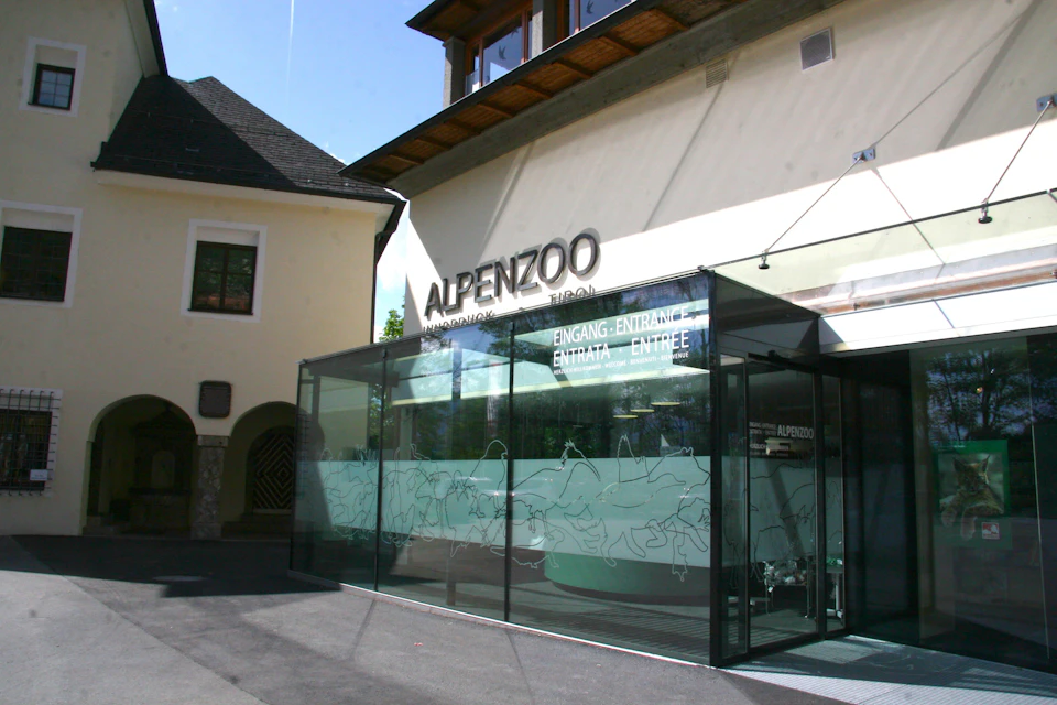 Glass entrance to Alpenzoo with multilingual "Entrance" signs on it, adjacent to a cream-colored building with a sloped roof.