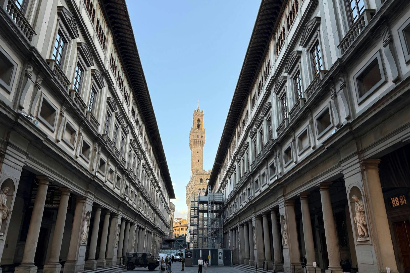 Tall, historic buildings line a narrow street, leading to a central tower under a clear sky. Few people walk in the distance.