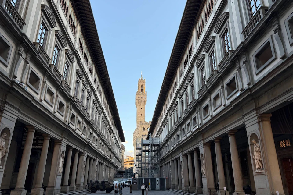 Tall, historic buildings line a narrow street, leading to a central tower under a clear sky. Few people walk in the distance.