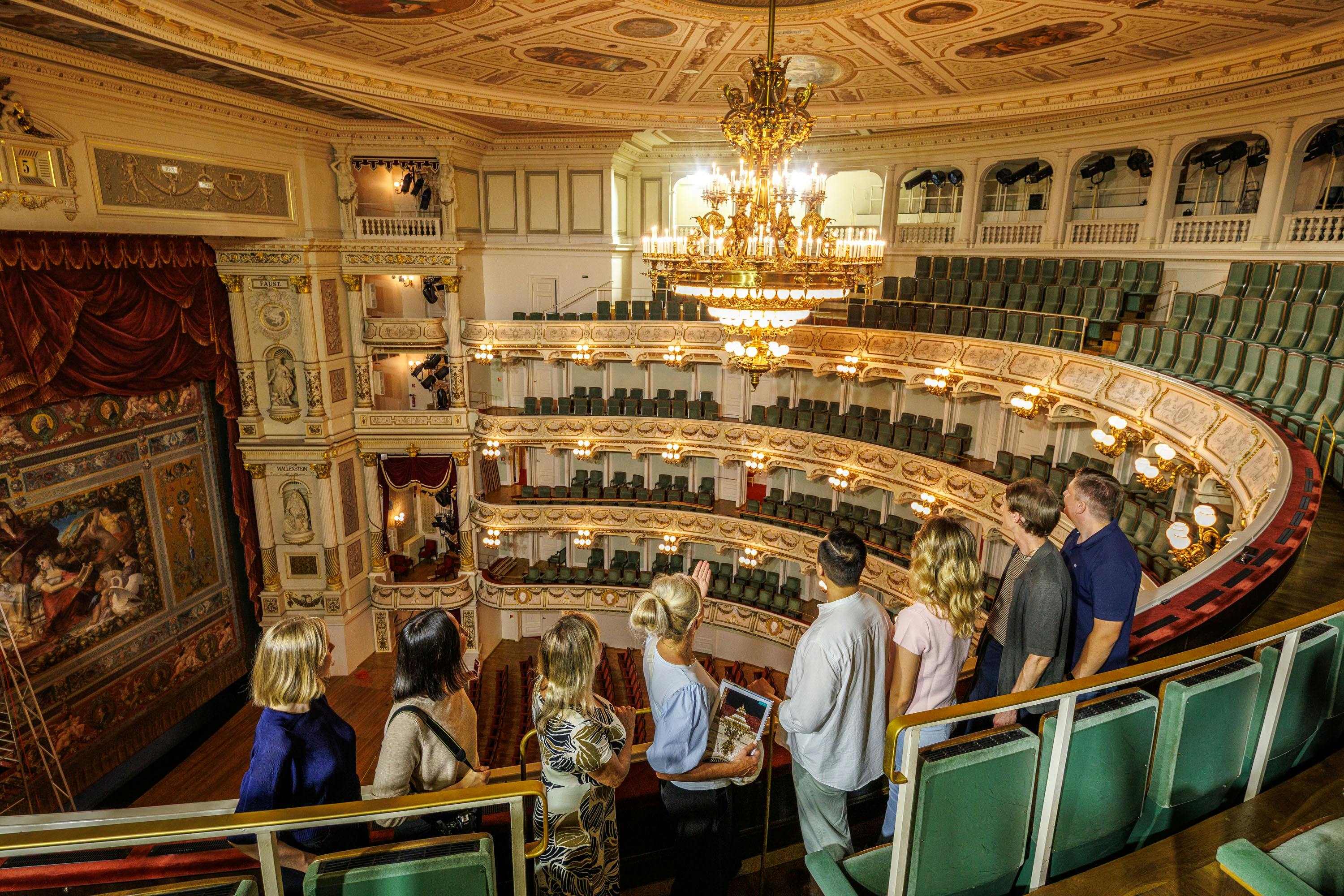 Um grupo de pessoas em pé na galeria de um teatro, a observar o interior ornamentado, com lustres e bancadas em camadas.