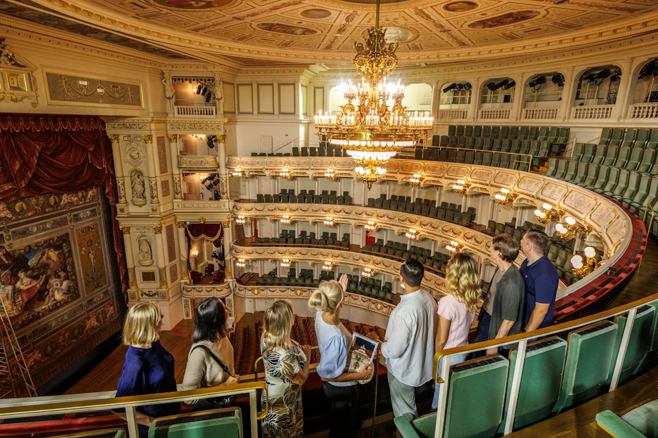A group of people standing in a theater balcony, observing the ornate interior with chandeliers and tiered seating.