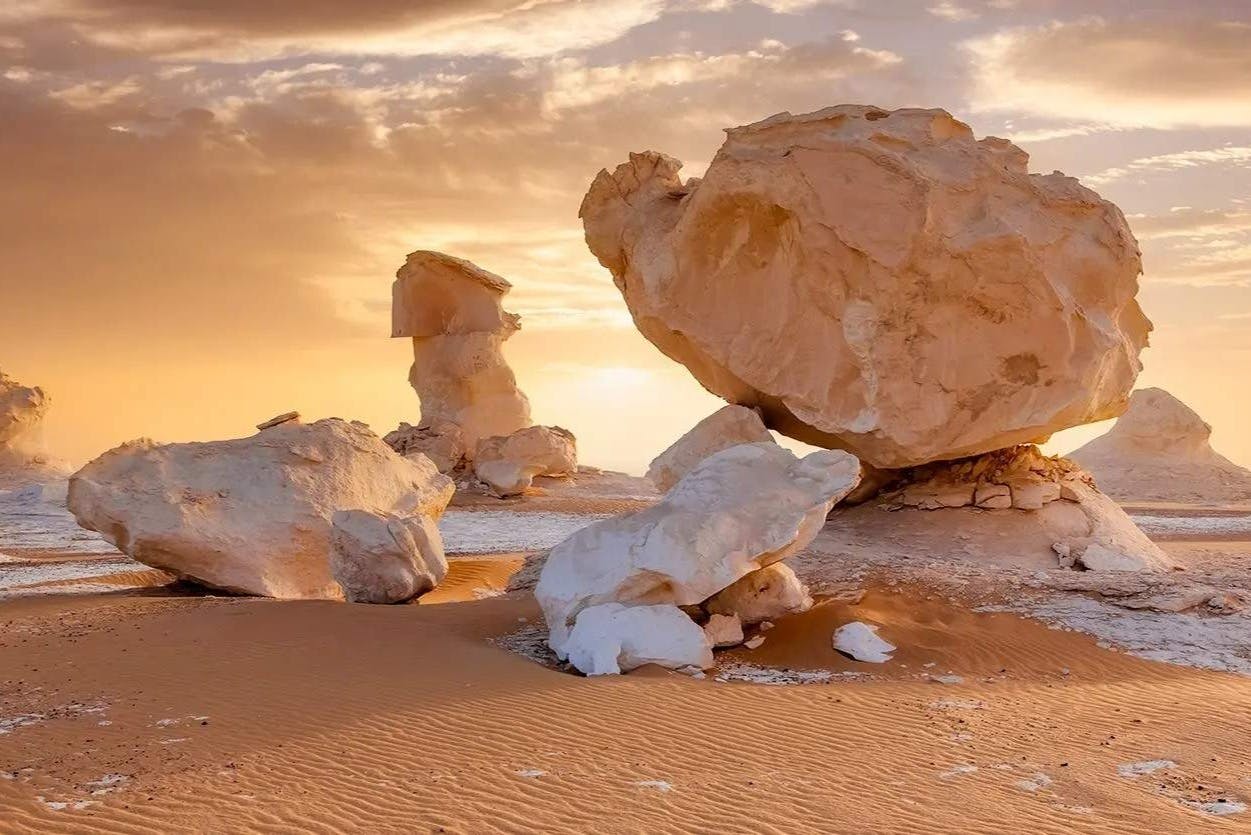 Large white and tan rock formations in a sandy desert landscape under a cloudy sky with warm lighting.