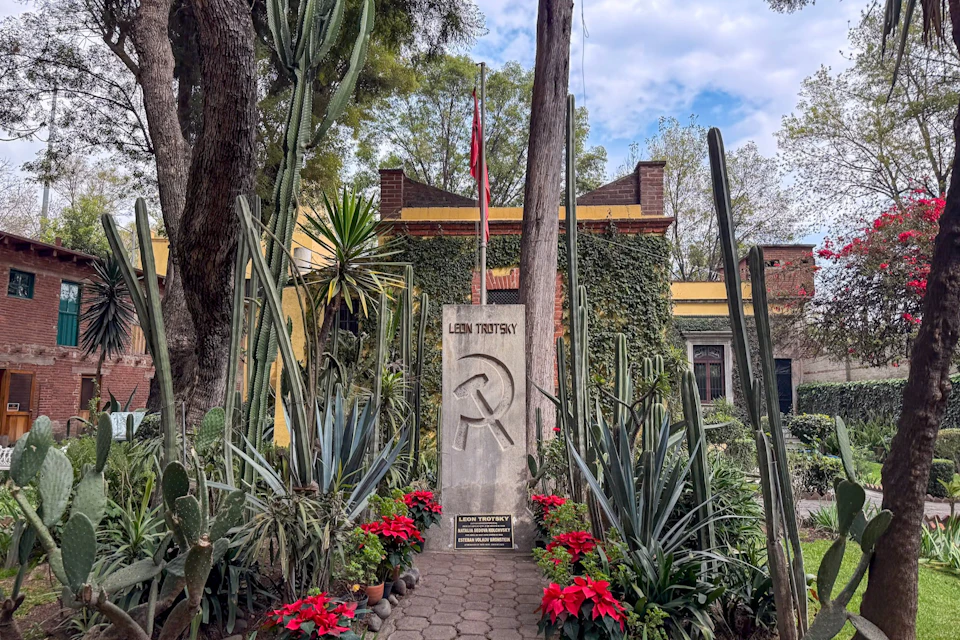 A stone monument with a hammer and sickle engraved, surrounded by plants, in front of a yellow building with flags and trees.
