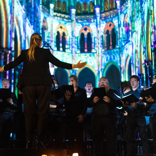 En dirigent leder en kör framför en färgglad, upplyst katedralbakgrund, med sångare som håller svarta mappar.