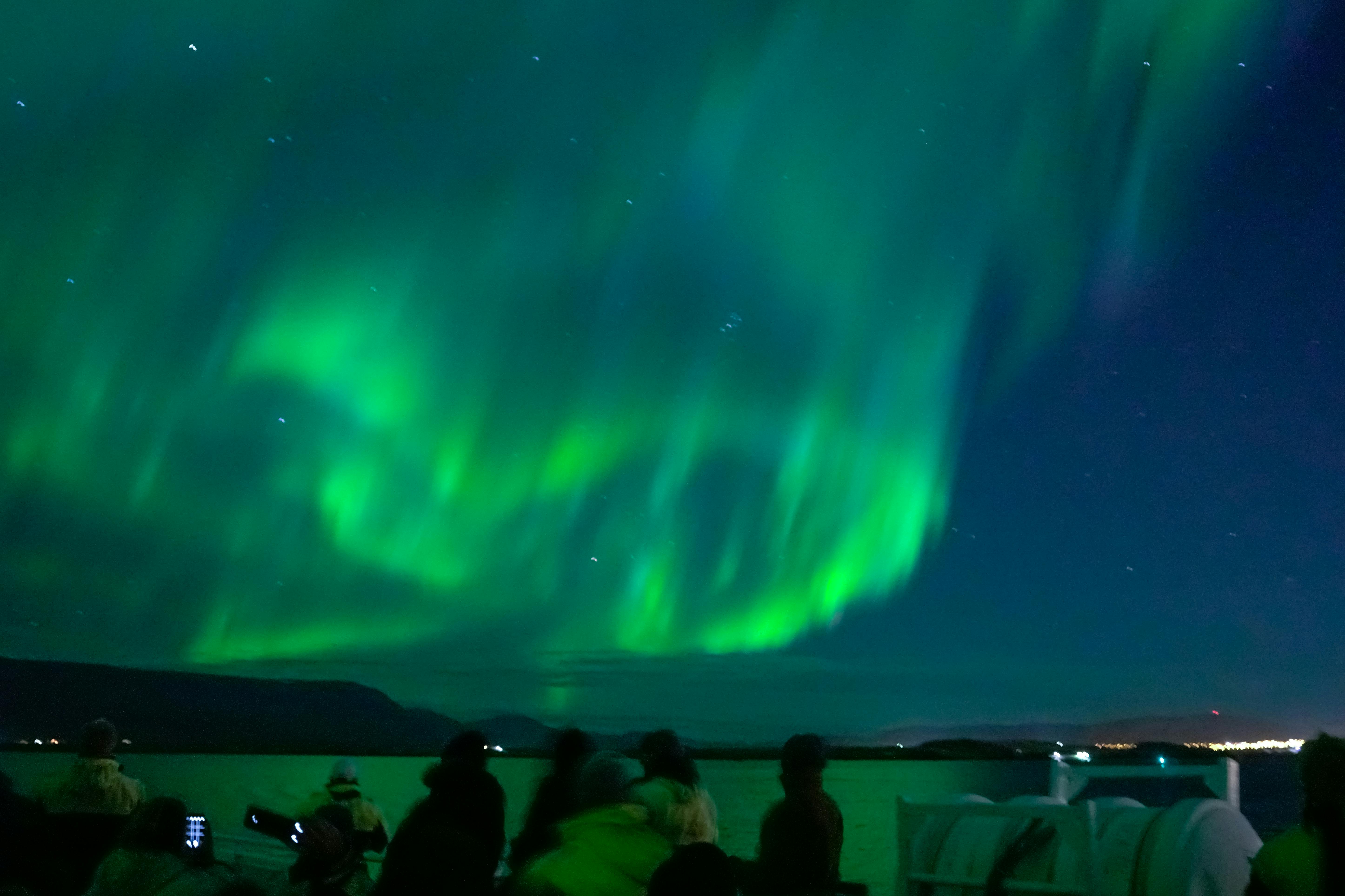People watching the vibrant green Northern Lights under a night sky. Dark silhouettes of mountains and water in the background.