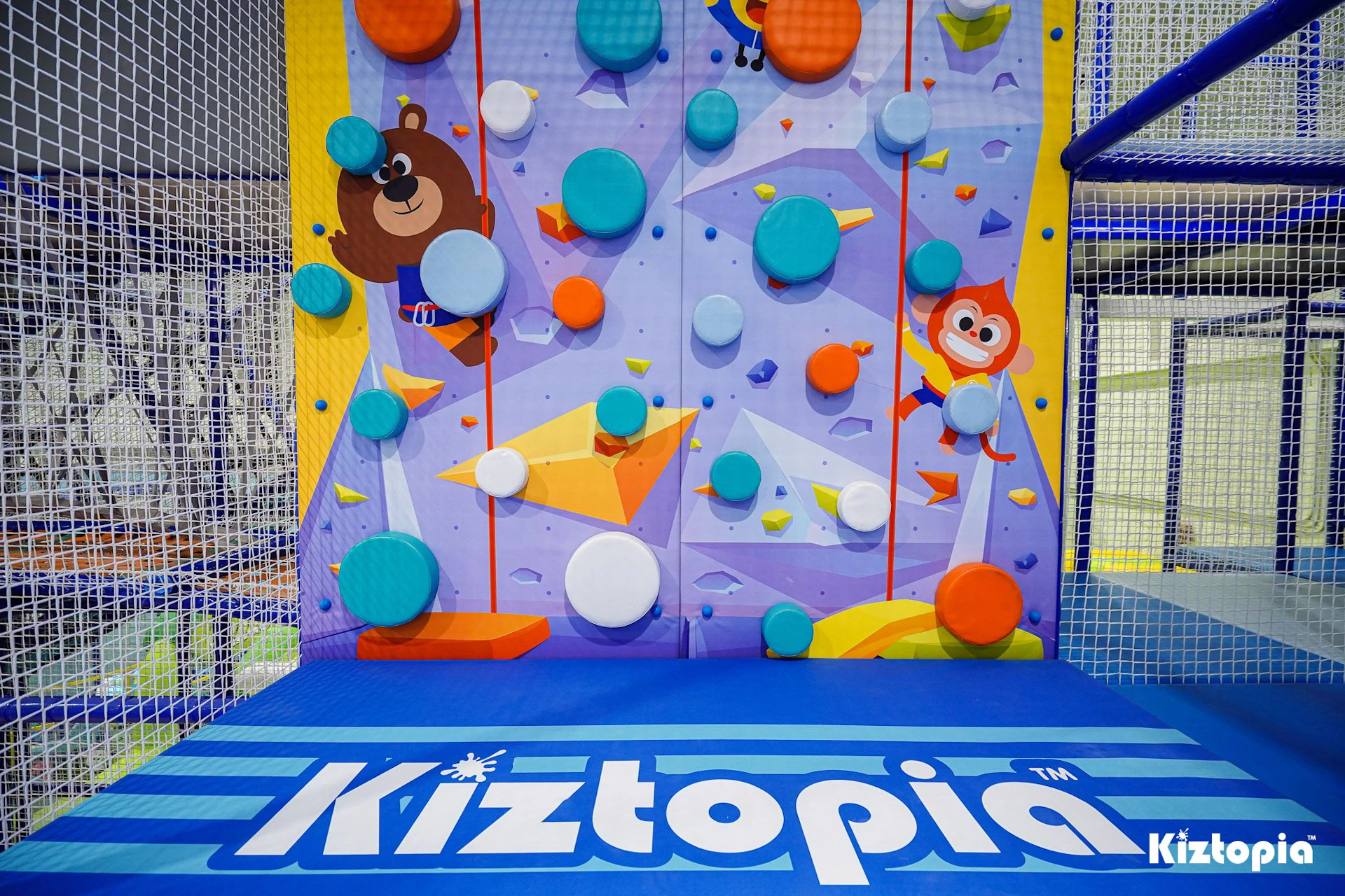 A colorful climbing wall with cartoon animal designs and padded holds, set in an indoor play area labeled Kiztopia.