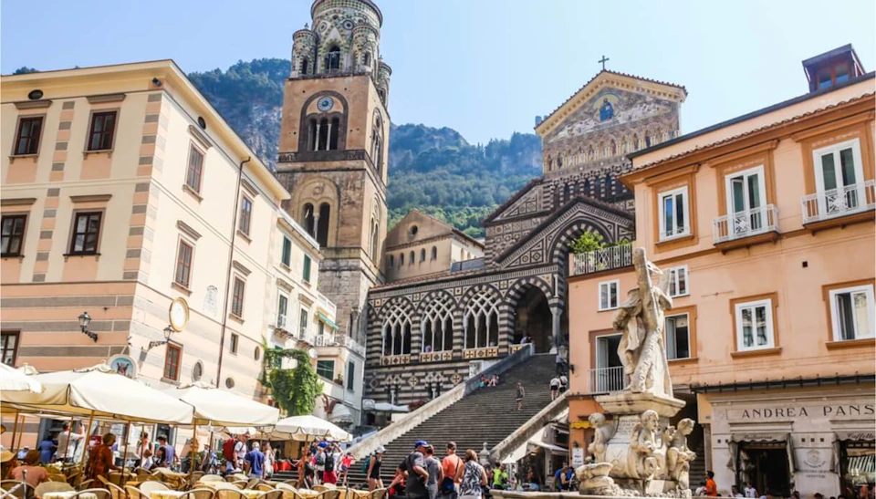 People walking and sitting near the Amalfi Cathedral, with its ornate facade and bell tower, surrounded by buildings and outdoor café tables.