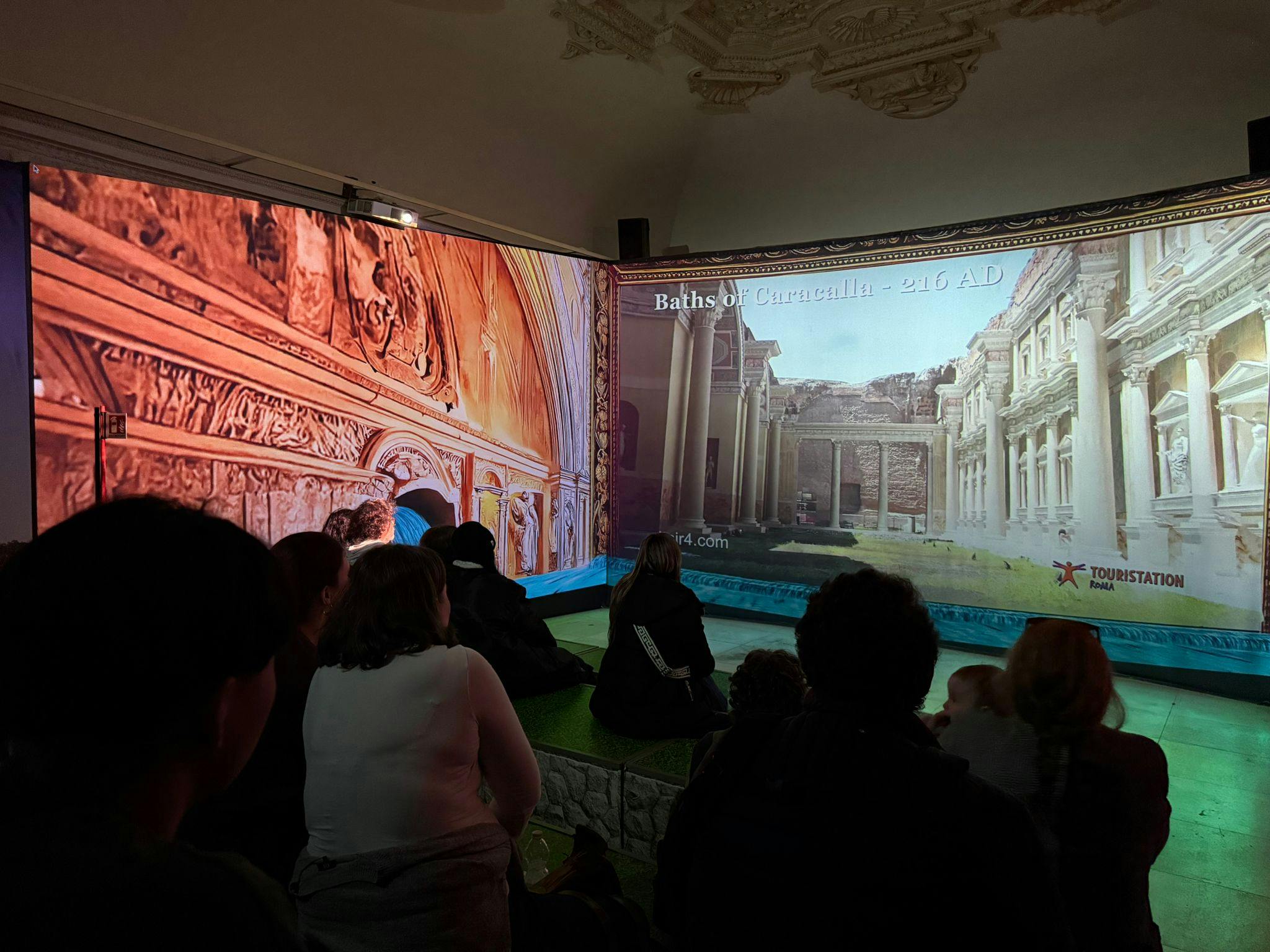 Un gruppo di persone sedute in una stanza guarda una proiezione di architettura storica su uno schermo. Il testo "Terme di Caracalla - 216 d.C." è visibile sul display.