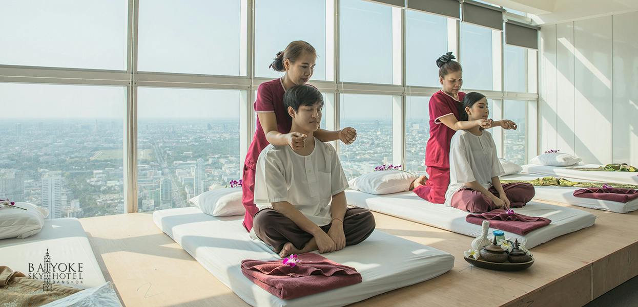 Two people receiving massages on floor mats in a well-lit room, with two practitioners in matching uniforms assisting them.