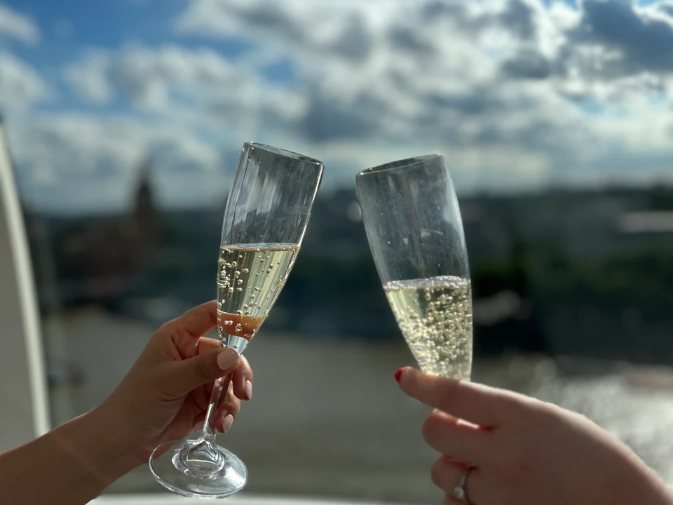 Two hands clinking champagne glasses against a blurred outdoor backdrop under a partly cloudy sky.