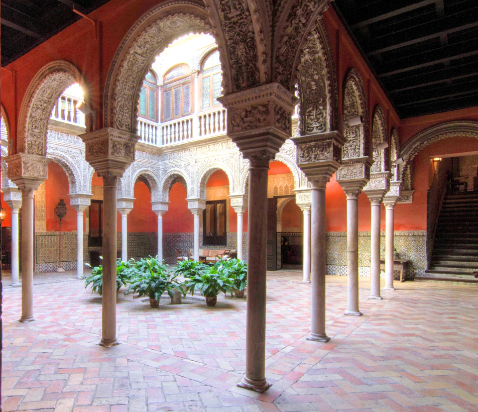A courtyard with ornate arches and columns, tiled walls, brick flooring, and a cluster of potted plants in the center.