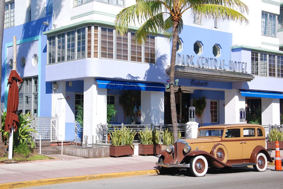 Vintage car parked in front of a pastel blue Art Deco building and palm tree in a sunny urban setting.