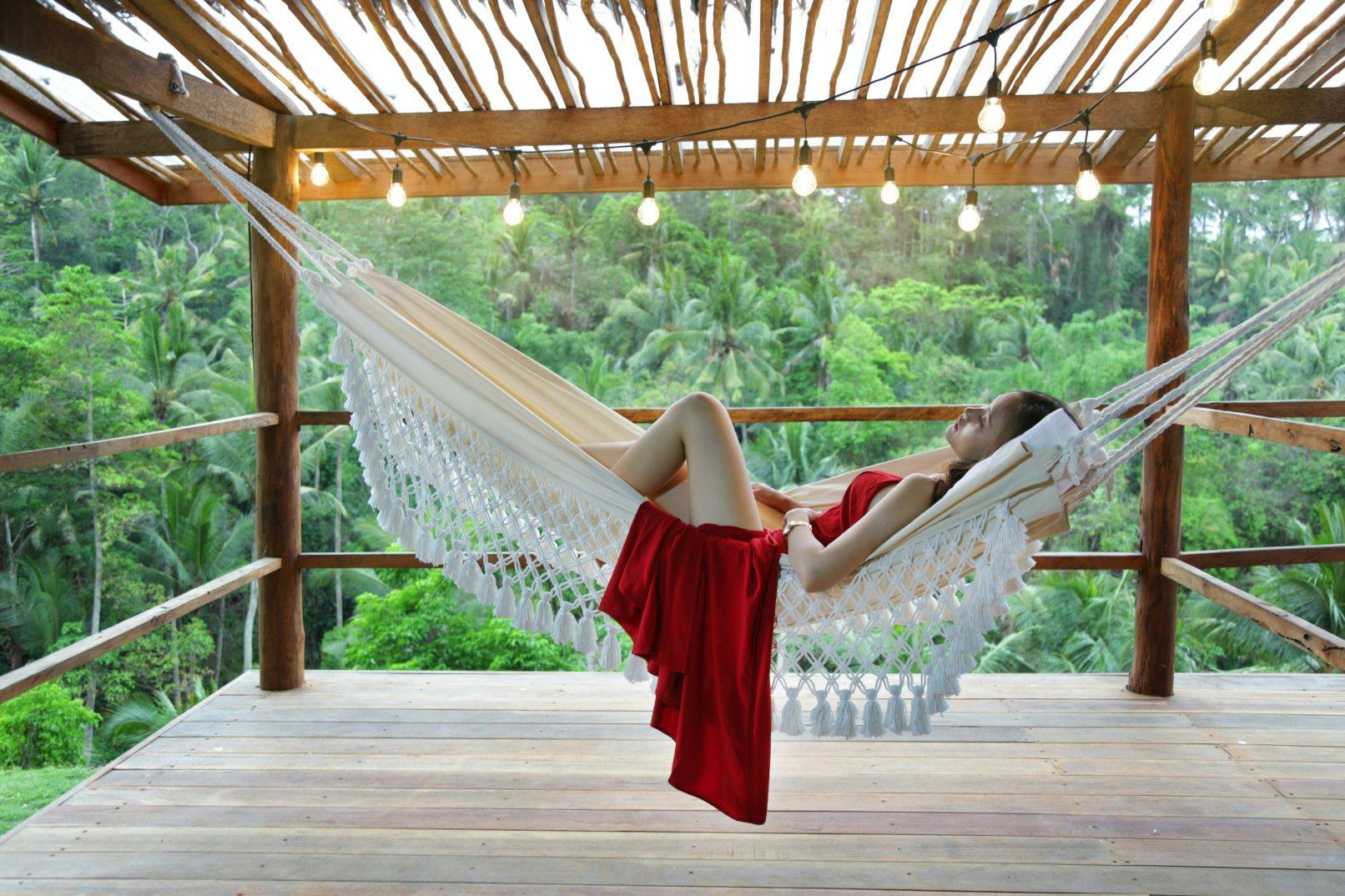 Une femme vêtue d'une robe rouge se détend dans un hamac sous une pergola en bois ornée de guirlandes lumineuses, avec vue sur une forêt verdoyante.