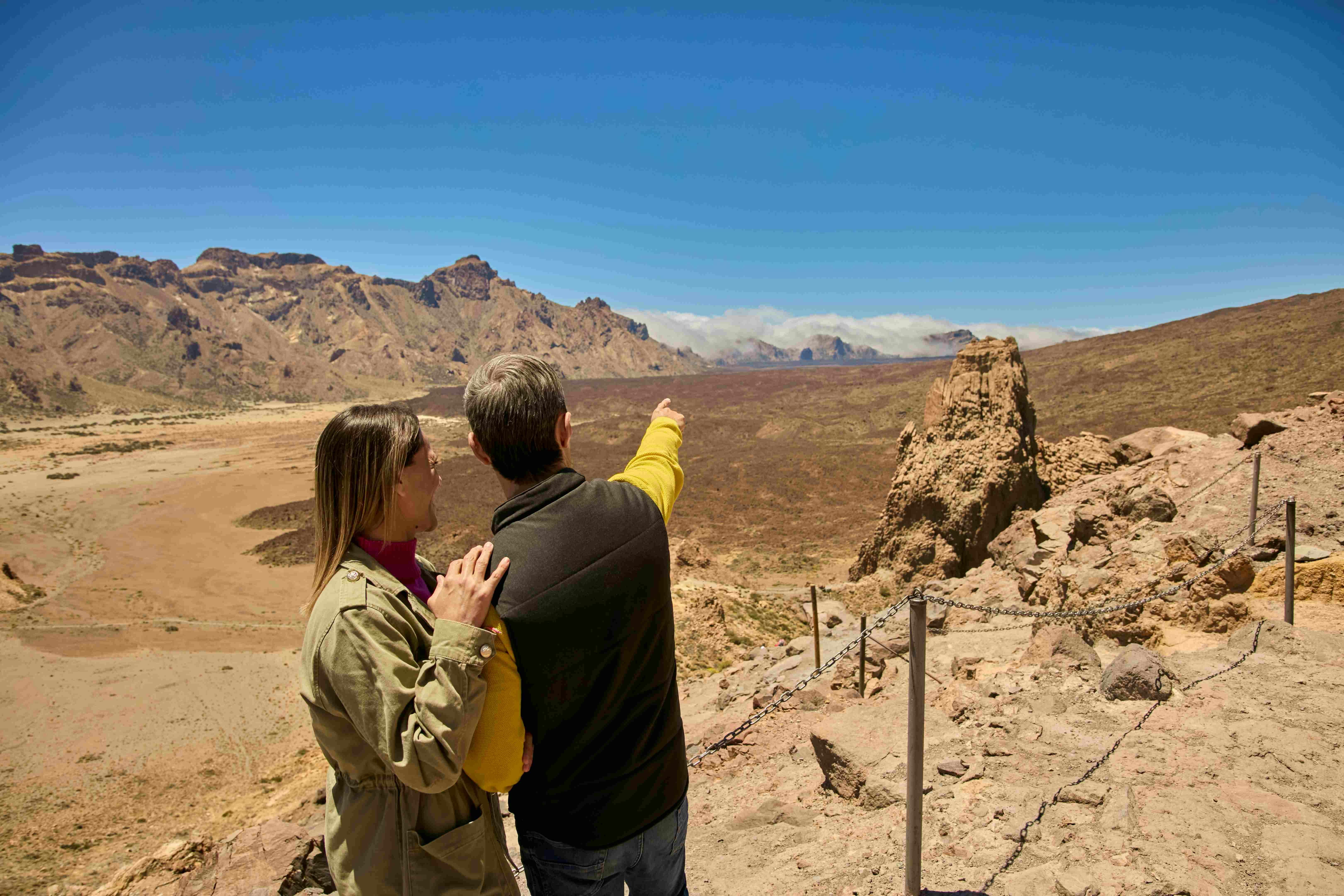 Two people standing on a rocky path, one pointing towards distant mountains under a clear blue sky.