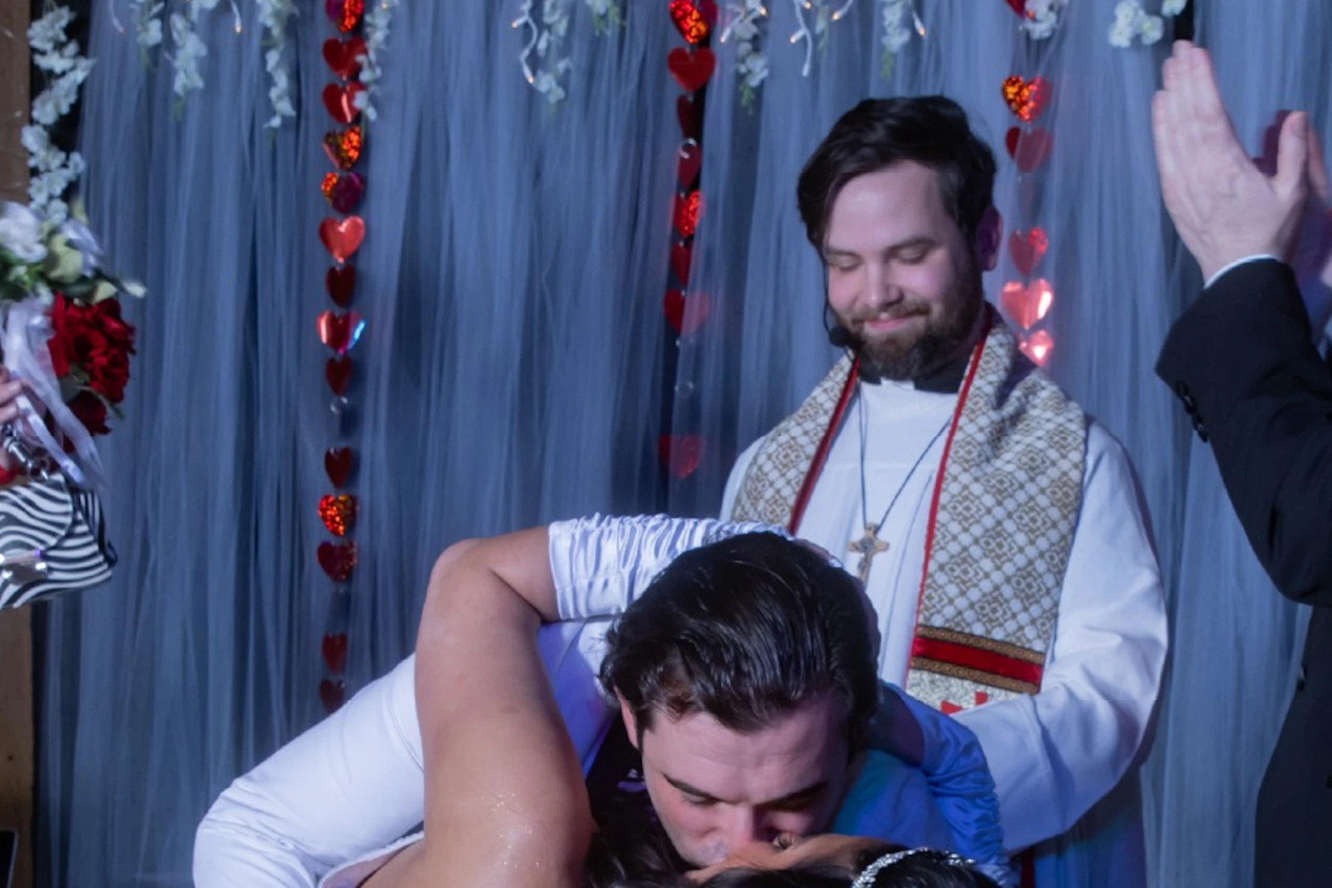 A bride and groom in a wedding ceremony kiss while others cheer. The setting includes decorations and a priest officiating.