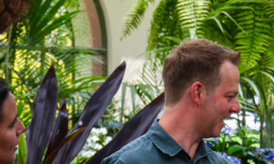 Two people, one in a blue shirt and one in a beige top, smiling and talking in a lush garden with hanging plants and flowers.