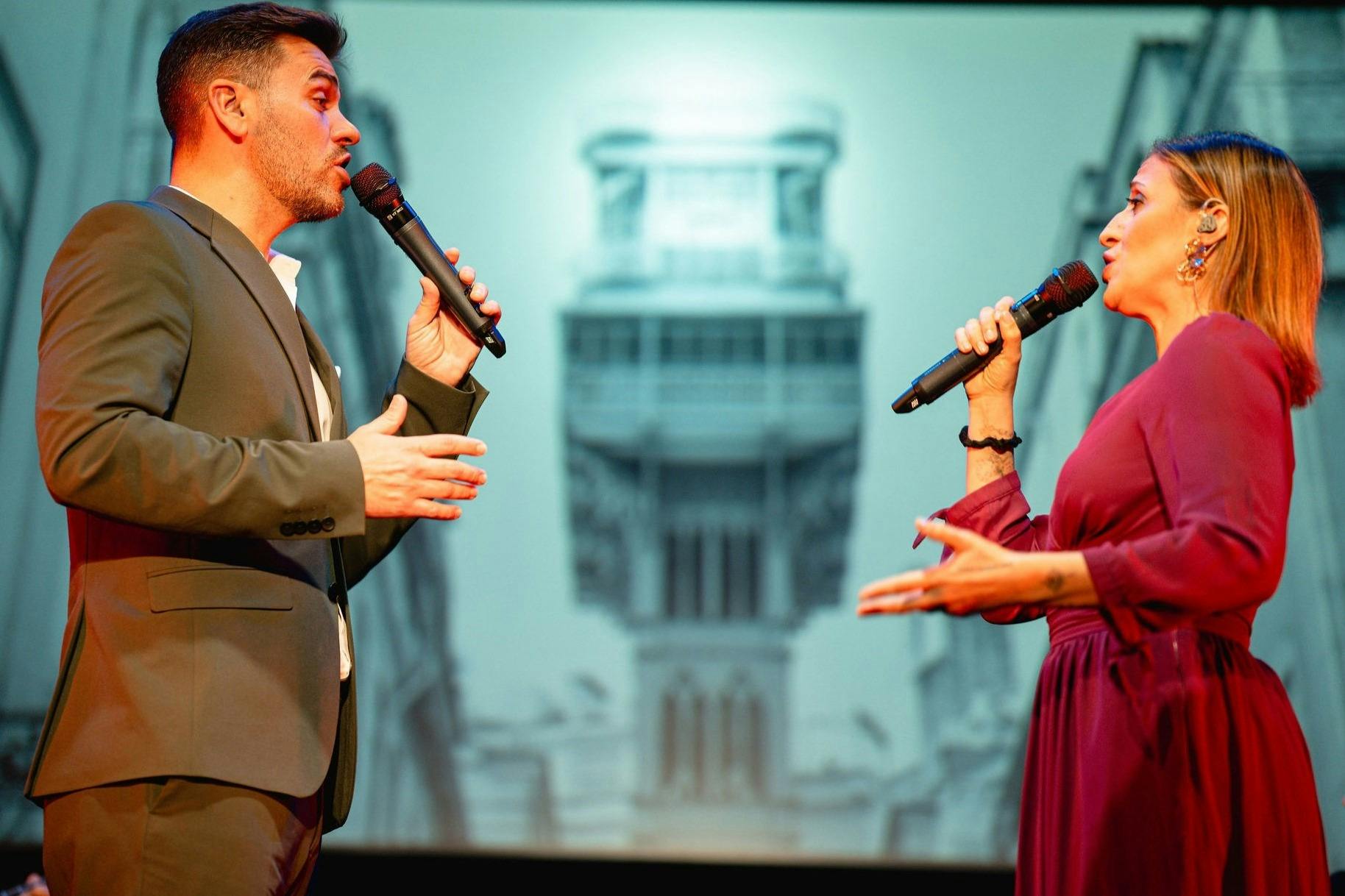 スーツを着た男性がステージ上でマイクに向かって話しかけています。背景には建物の映像が映し出されています。