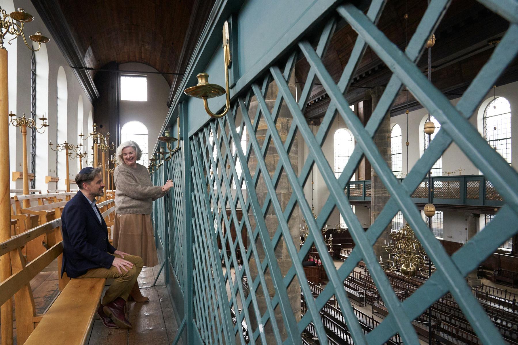 Deux personnes discutent sur un balcon en bois à l'intérieur d'une église, avec de grandes fenêtres et des chandeliers en laiton ornés le long des murs.