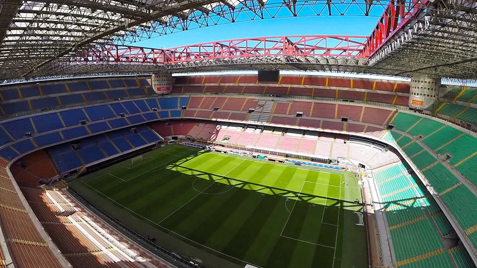 Empty football stadium with green field, multi-colored seats, and a large red roof structure. Bright sunlight casts shadows.