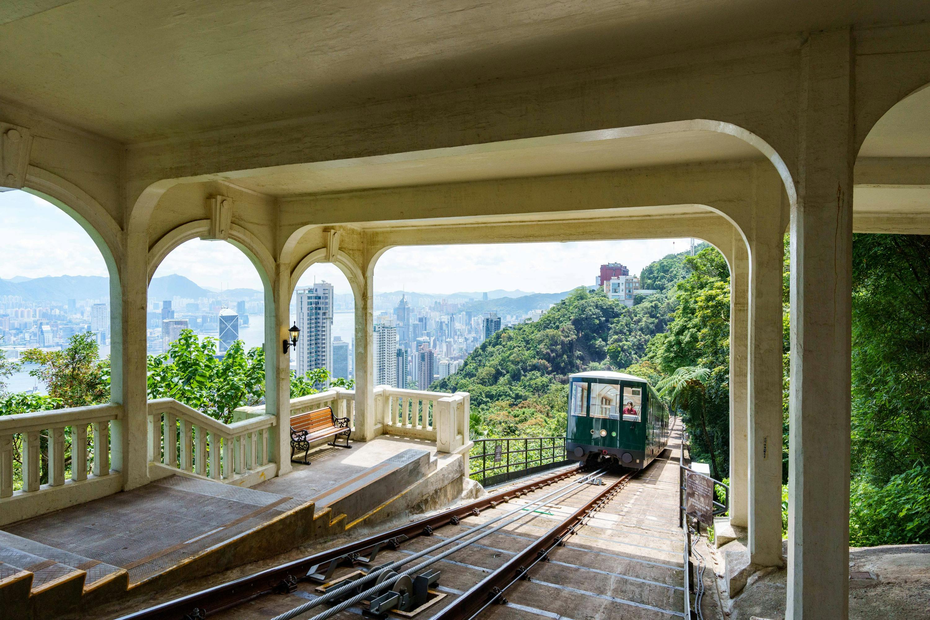 A tram at a station on a hill with a view of a city skyline and forested mountains in the background, framed by an archway.