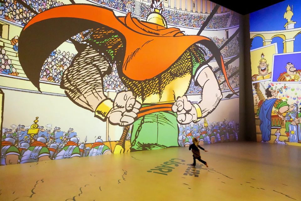 A child runs in front of a large projected comic-style image of a gladiator in an arena filled with spectators.