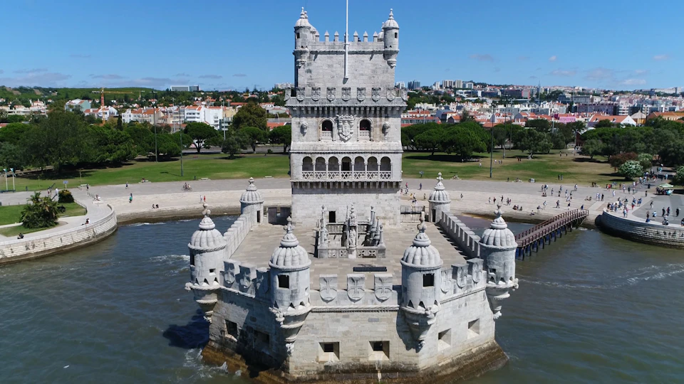 Historic stone tower with battlements and turrets, surrounded by water and a park area. City and blue sky in the background.