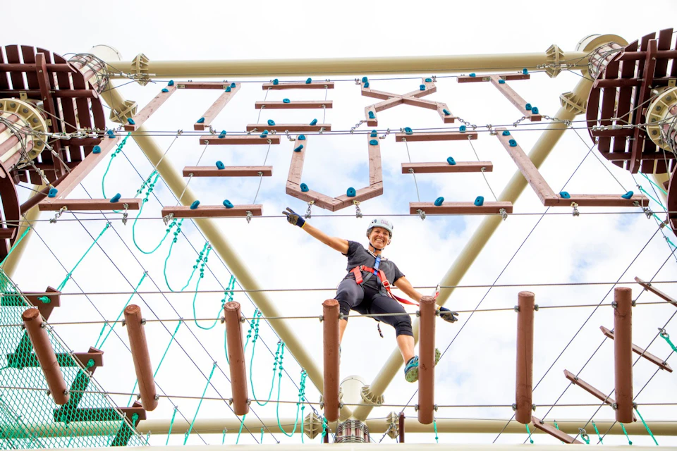 Person in harness navigating an elevated rope course, smiling with one arm outstretched. Structures and ropes are visible.