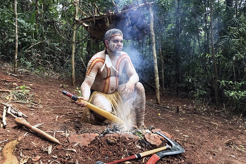 바디 페인트와 전통 복장을 한 남성이 나무 도구를 들고 숲 속 작은 불 근처에서 웅크리고 있습니다.