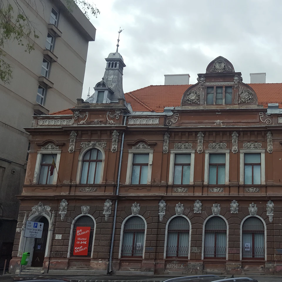 Ornate brown building with decorative stonework, arched windows, and a red tiled roof. Adjacent to a gray multi-story building.