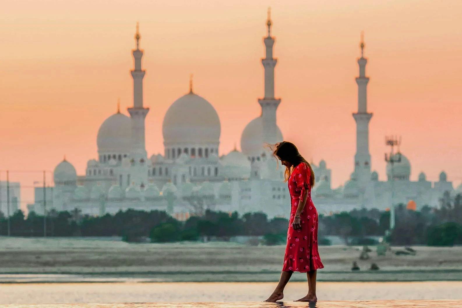 A person in a red dress walks near water with a mosque featuring domes and minarets in the background during sunset.