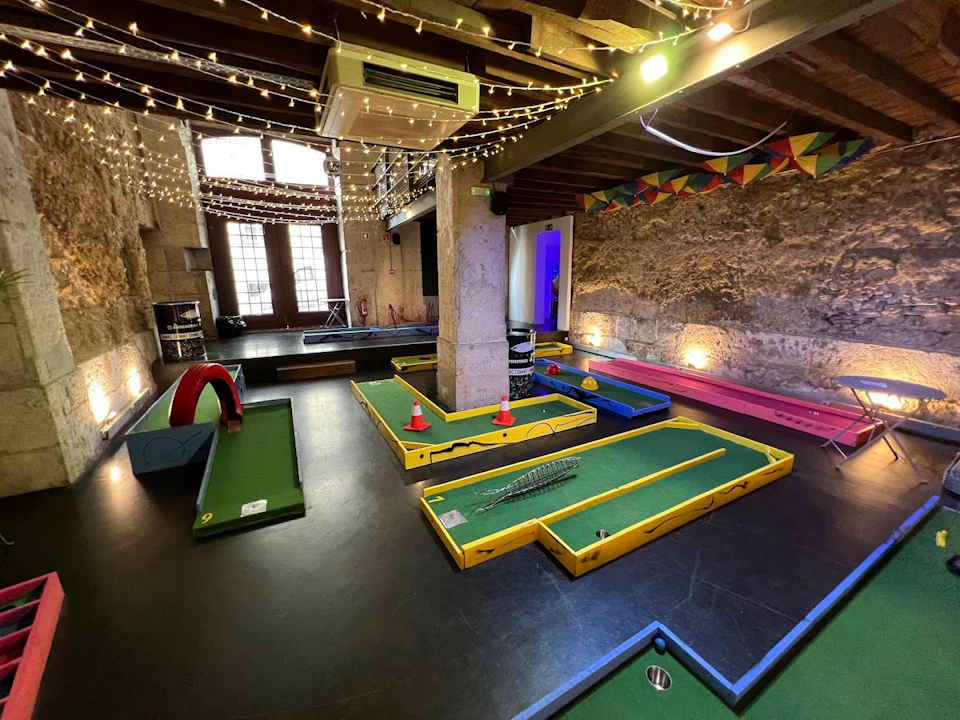 Indoor mini-golf course with various obstacles, string lights on ceiling, exposed stone walls, and a colorful bunting decoration.