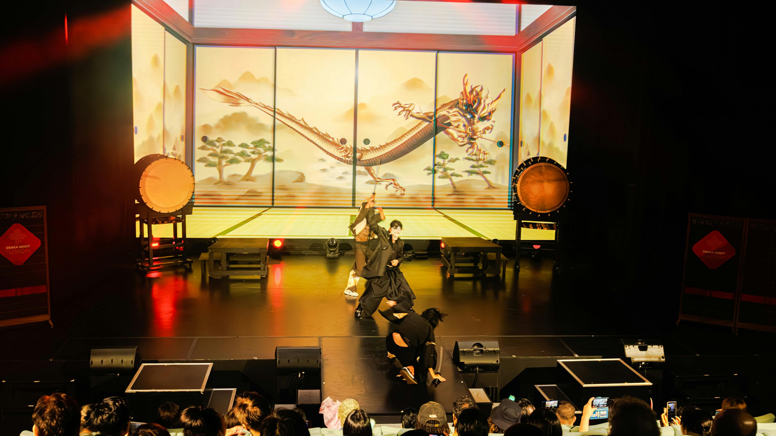 Performers on a stage depicting a traditional Japanese setting with a dragon mural and large taiko drums in the background.