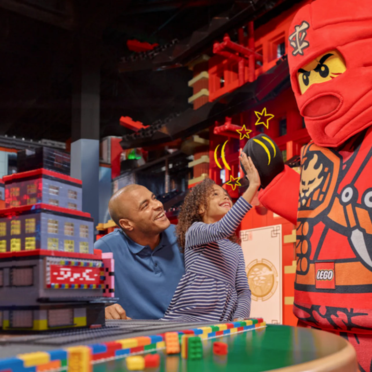 Child high-fives a person in a LEGO ninja costume, with a smiling adult nearby. LEGO buildings are in the foreground.