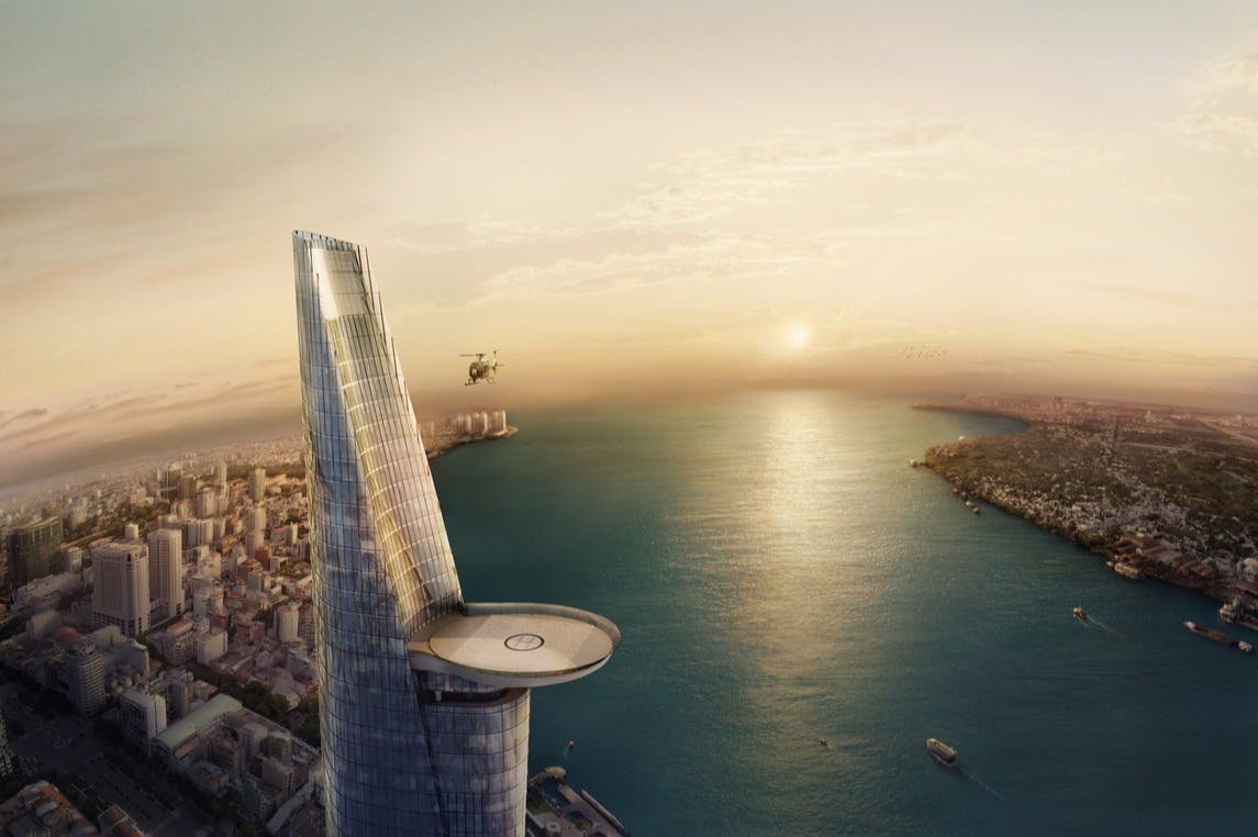 Helicopter landing on a helipad atop a tall skyscraper by a waterfront at sunset. Urban landscape and water visible below.