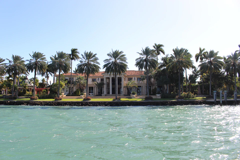 A large waterfront villa with palm trees and a red-tiled roof, seen across turquoise water under a clear blue sky.