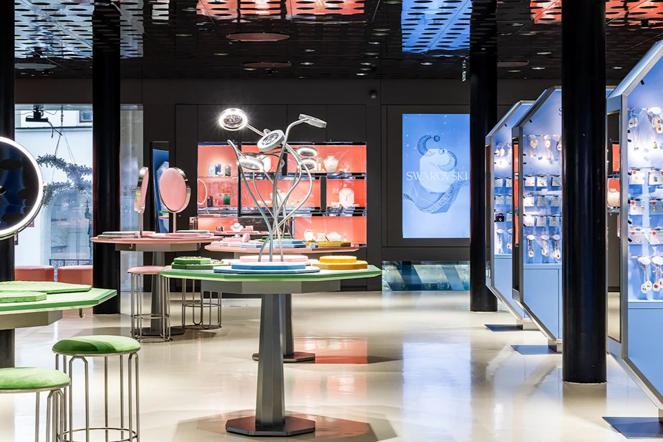 Modern store interior with various colorful display tables and shelves. One shelf features a "Swarovski" sign and jewelry.