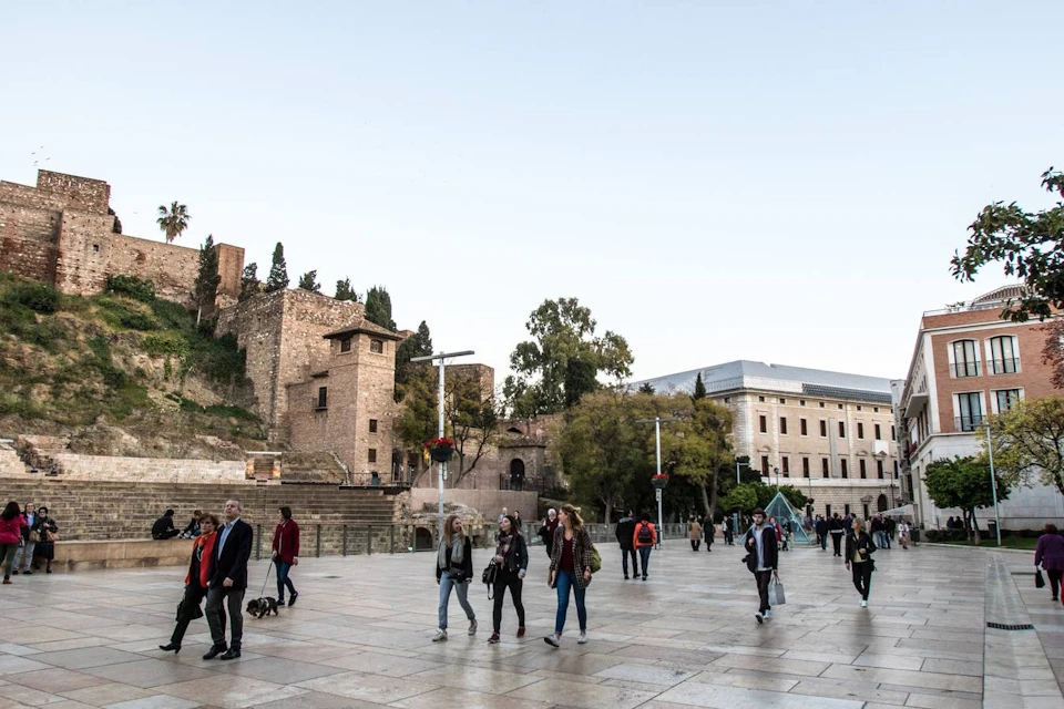 People walking in a plaza near historic stone buildings and a modern structure, with trees and a clear sky in the background.