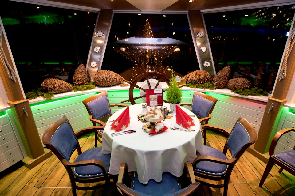 A festive, round dining table set for four with red napkins and holiday decor. A ship's wheel and large window decorated with lights in the background.