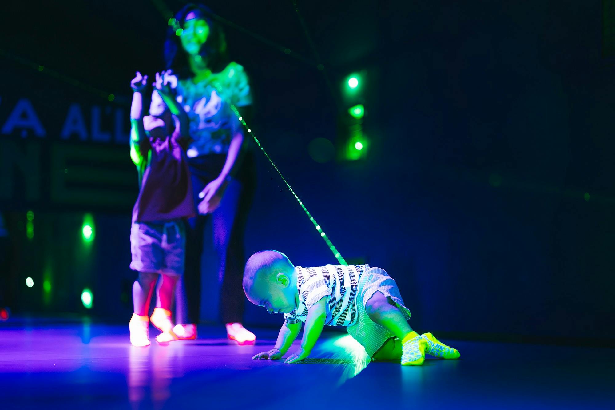 En baby som kryper på ett golv under färgglada ljus, med ett barn och en vuxen i bakgrunden, i ett svagt upplyst område.