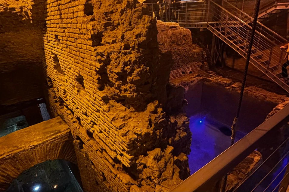 Ancient brick walls lit by warm lighting, with a blue-lit area below and modern railings in the background.
