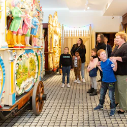 En grupp människor, inklusive barn, observerar en färgglad, dekorativ orgelutställning i en inomhus museumsliknande miljö.