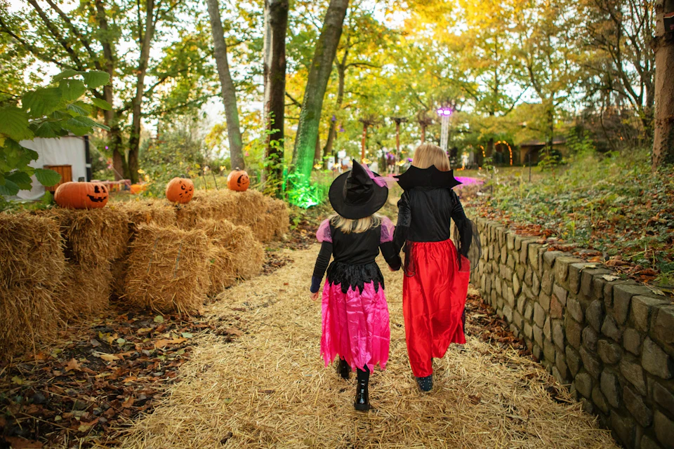 Two children in Halloween costumes walk along a straw-covered path in a wooded area decorated with pumpkins.
