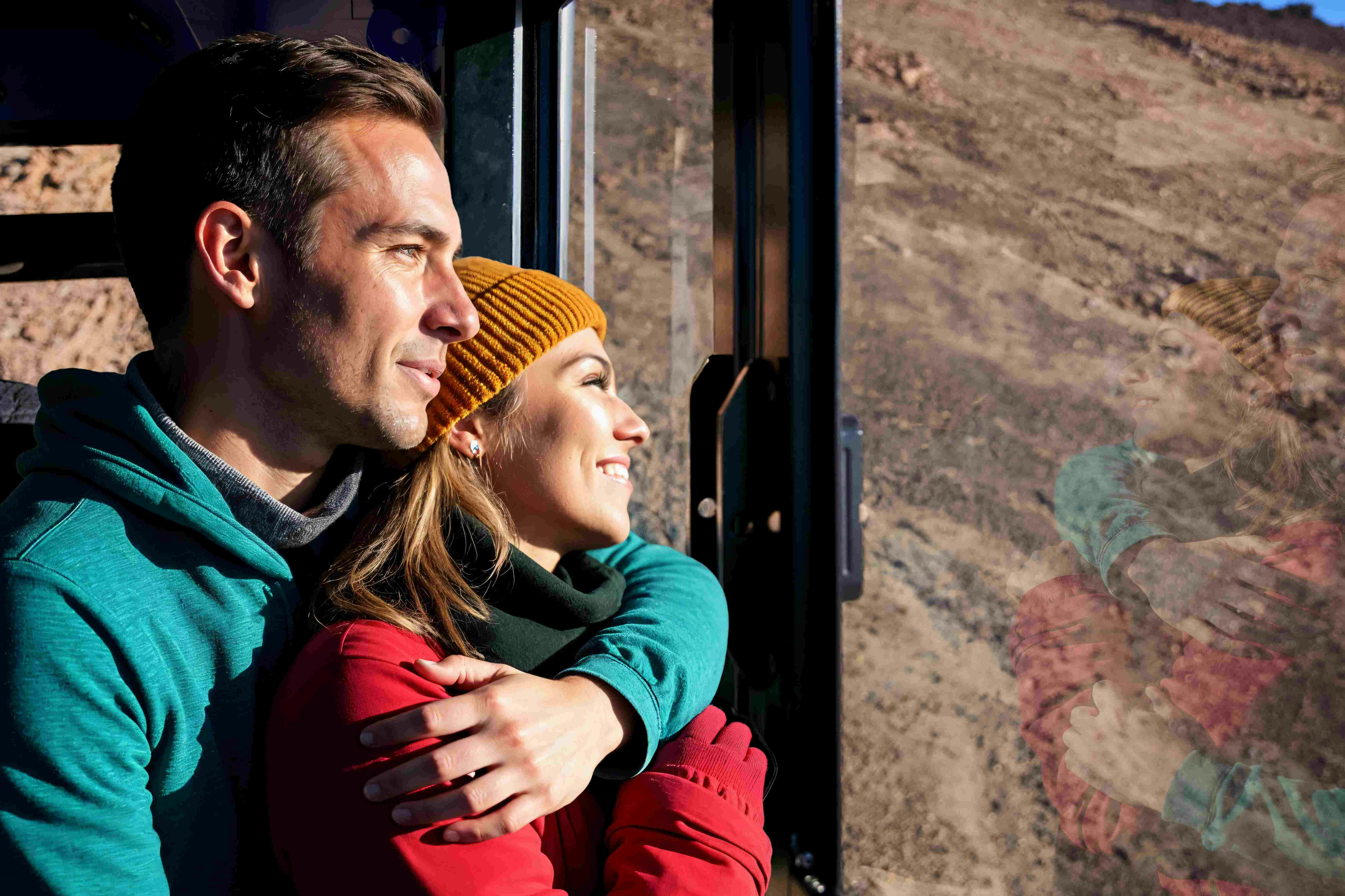 一对穿着保暖衣服的夫妇，男人从后面拥抱女人，透过窗户看着山景。