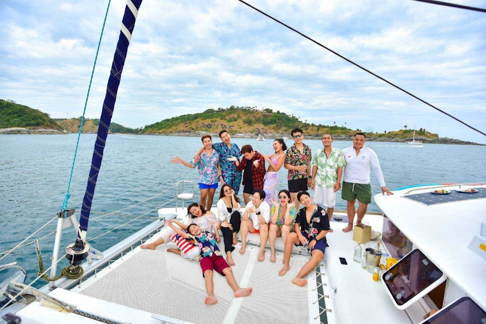 A group of people enjoying themselves on a boat, with an island and sailboats in the background under a partly cloudy sky.