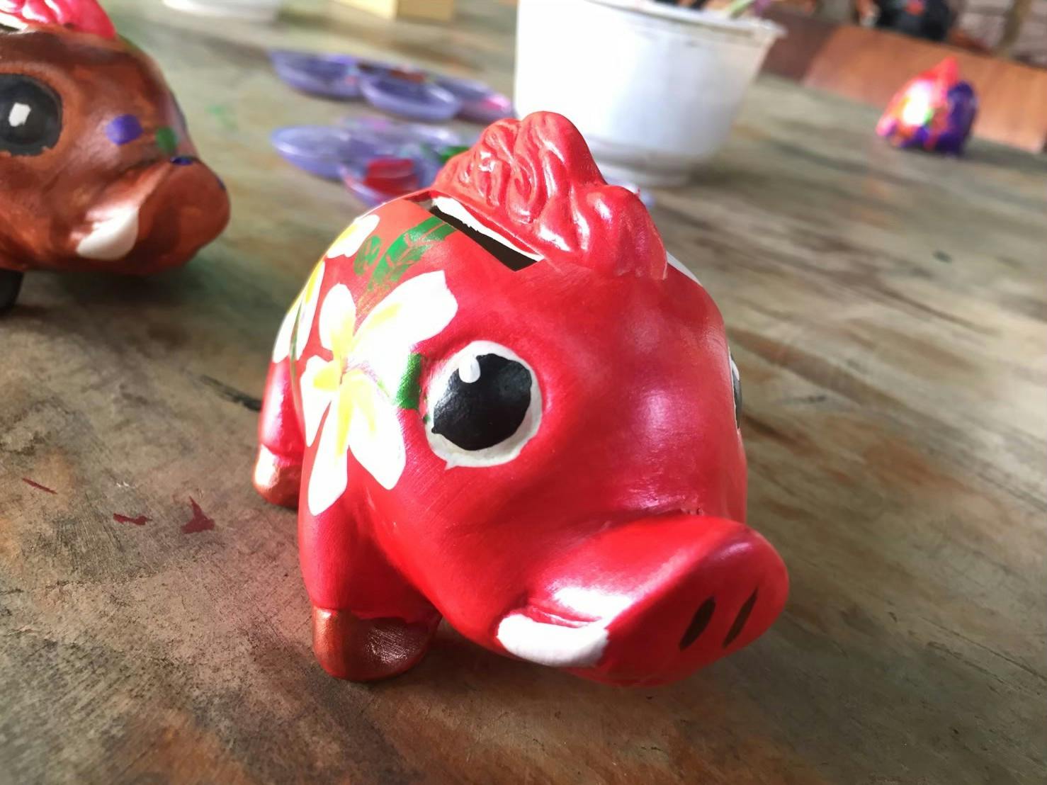 A red, painted piggy bank with floral designs, sitting on a wooden table with art supplies in the background.
