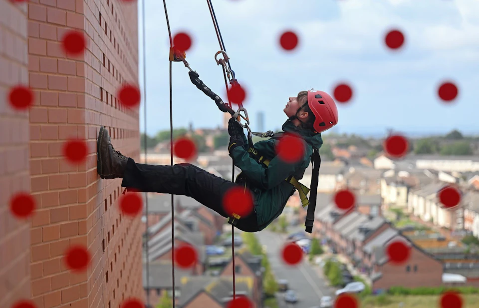 Person in safety gear rappelling down a brick wall with a red helmet and harness. Red dots overlay the image. Houses visible below.