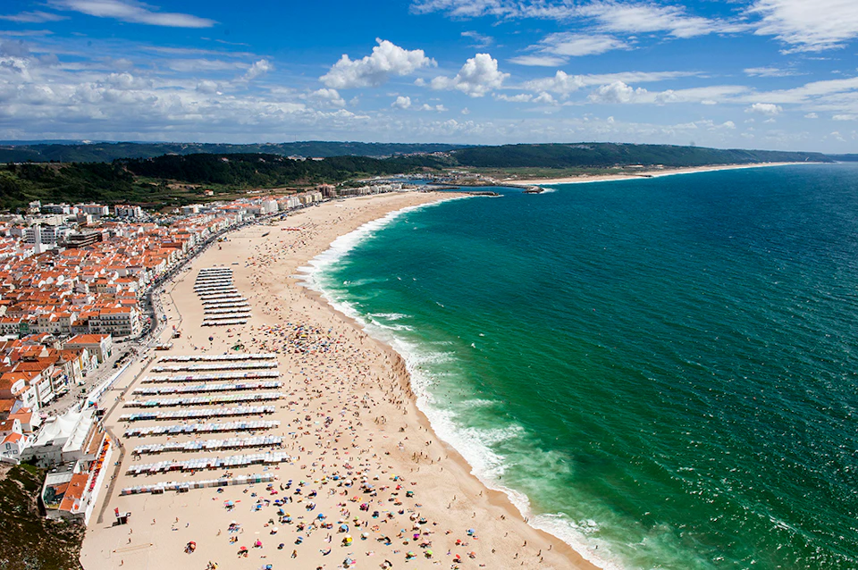 Aerial view of a crowded beach with rows of umbrellas, people sunbathing, and swimming in the sea, with buildings and hills in the background.