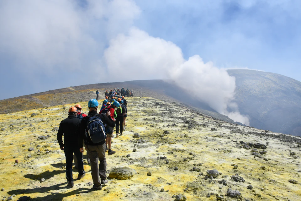 A group of people wearing helmets and backpacks hike along a rocky, yellowish terrain with smoke rising in the background.